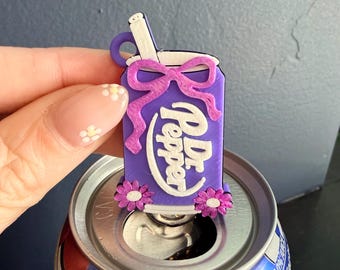 May include: A purple Dr. Pepper can-shaped charm with a white straw and a purple bow. The charm has the Dr. Pepper logo in white and is decorated with small pink flowers. The charm is placed on top of a Dr. Pepper can.