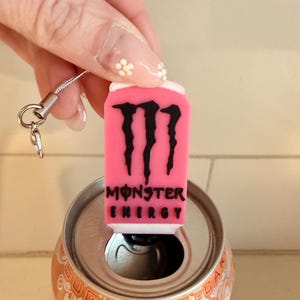 May include: A pink Monster Energy can charm with a black logo and text, held above an open orange soda can. The charm has a silver lanyard attached.