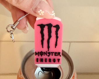 May include: A pink Monster Energy can charm with a black logo and text, held above an open orange soda can. The charm has a silver lanyard attached.