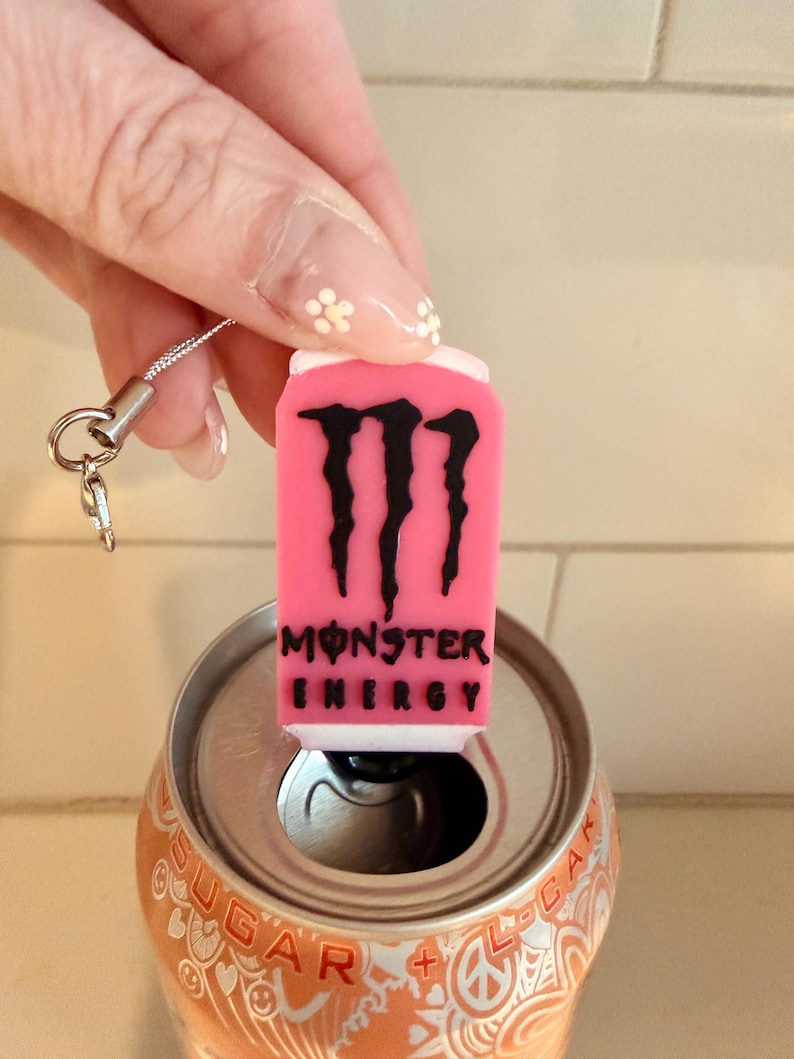 May include: A pink Monster Energy can charm with a black logo and text, held above an open orange soda can. The charm has a silver lanyard attached.