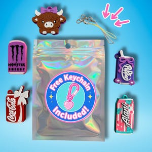 May include: A collection of colorful charms, including a brown Highland cow, a Monster Energy drink can, a Coca-Cola can, a Dr. Pepper bottle, and an Alani Nu can. A clear bag with a sticker that says "Free Keychain Included!" is in the center. A yellow lanyard is also included.