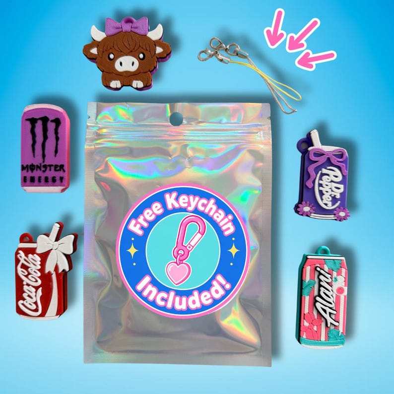 May include: A collection of colorful charms, including a brown Highland cow, a Monster Energy drink can, a Coca-Cola can, a Dr. Pepper bottle, and an Alani Nu can. A clear bag with a sticker that says "Free Keychain Included!" is in the center. A yellow lanyard is also included.