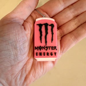 May include: A pink pendant shaped like a Monster Energy drink can, with the brand's logo and text in black. The pendant is suspended from a silver-colored chain. The can design has a white top and bottom.