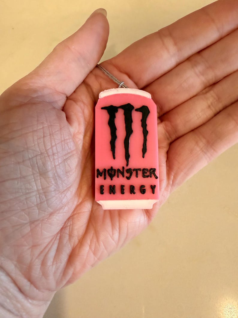 May include: A pink pendant shaped like a Monster Energy drink can, with the brand's logo and text in black. The pendant is suspended from a silver-colored chain. The can design has a white top and bottom.