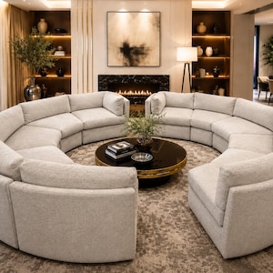 May include: A large, curved sectional sofa in a light gray fabric dominates the room. A round, black coffee table with a gold rim sits in the center. The room features a fireplace, artwork, and a neutral color palette.