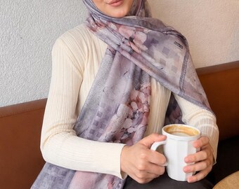 Op de afbeelding: Een crèmekleurige geribbelde top met lange mouwen gecombineerd met een donkerbruine rok en een lavendelkleurige hijab met een bloemenpatroon en een geblokte rand. De hijab is over het hoofd en de schouders gedrapeerd en een witte mok wordt vastgehouden.