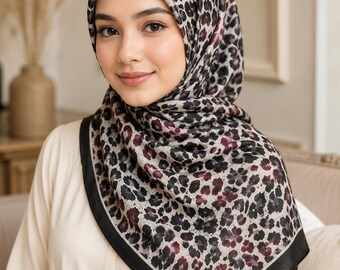 Op de afbeelding: Een vierkante hoofddoek met een gedetailleerd luipaardprintpatroon in tinten zwart, grijs en diep bordeauxrood. De sjaal heeft een effen zwarte rand langs de zijkanten en is in een traditionele hijab-stijl om het hoofd en de nek gedrapeerd.