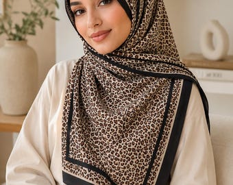 Op de afbeelding: Een vierkante hoofddoek met een bruin en zwart luipaardprintpatroon en een effen zwarte rand. De stof is over het hoofd en de schouders gedrapeerd en toont een herhalend dierenprintontwerp dat wordt omlijst door een duidelijke zwarte bies.