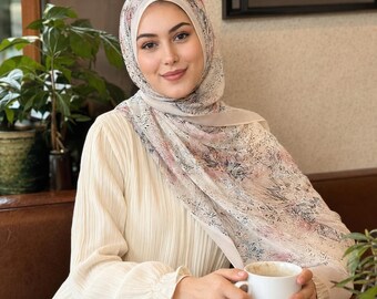Op de afbeelding: Een crèmekleurige blouse met lange mouwen en plooien, gecombineerd met een lichtgewicht hoofddoek met een delicaat bloemenpatroon in zachte tinten roze, grijs en zwart. De hoofddoek is elegant gedrapeerd rond het hoofd en de schouders.