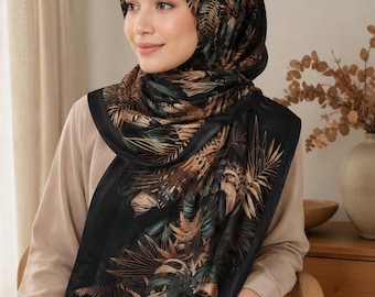 Op de afbeelding: Een rechthoekige hoofddoek met een zwarte basis en een gedetailleerde botanische print van palmbladeren en tropisch gebladerte in tinten brons, koper en gedempt blauwgroen. De stof heeft een gladde, satijnachtige afwerking en is rond het hoofd en de nek gedrapeerd.