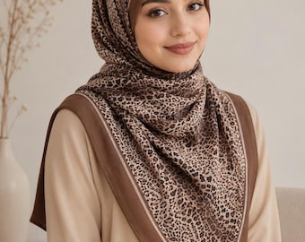 Op de afbeelding: Een vierkante hoofddoek met een bruin en zwart luipaardpatroon in het midden, omlijst door een effen donkerbruine rand met een dunne lichtbeige lijn. De sjaal is over het hoofd en de schouders gedrapeerd en gecombineerd met een beige top.