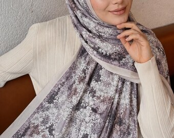 Op de afbeelding: Een rechthoekige hijab met een gedetailleerd bloemen- en abstract patroon in tinten grijs, wit en gedempt paars. De stof heeft een zachte textuur met een effen lichtgrijze rand langs de zijkanten, elegant gedrapeerd over het hoofd en de schouders.