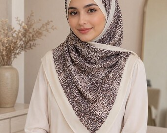 Op de afbeelding: Een vierkante hoofddoek met een bruin en zwart luipaardpatroon op een lichtbeige achtergrond, afgewerkt met een effen crèmekleurige rand. De sjaal is over het hoofd en de schouders gedrapeerd en omlijst het gezicht met zachte, neutrale tinten.