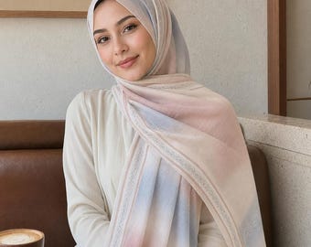 Op de afbeelding: Een lichtgewicht hijab met een zacht kleurverloop van pastelroze, blauwe en crèmetinten. De stof heeft een delicaat, ingewikkeld randpatroon in een subtiele zilveren of lichtgrijze tint, dat de randen van de rechthoekige hoofdbedekking omlijst.