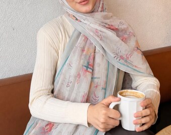 Op de afbeelding: Een lichtgrijze hijab met een delicaat bloemenpatroon in roze, oranje en donkerrode tinten, geaccentueerd met een blauwgroene rand. De hijab wordt gedragen over een crèmekleurige geribbelde top met lange mouwen, gecombineerd met een donkerbruine rok en een witte mok.