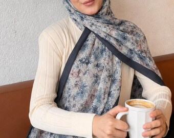 Op de afbeelding: Een hijab met bloemenprint in tinten blauw, grijs en gedempt roze met een effen donkerblauwe rand, gedragen over een crèmekleurige geribbelde top met lange mouwen en een donkerbruine rok, terwijl een witte keramische mok met koffie wordt vastgehouden.