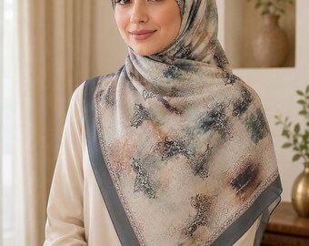 Op de afbeelding: Een vierkante hoofddoek met een beige basis en een veelkleurig abstract patroon van donkerblauwe, groene en roestkleurige ornamenten. De sjaal heeft een effen donkergrijze rand langs de zijkanten en wordt gedrapeerd over hoofd en schouders.