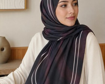 Op de afbeelding: Een rechthoekige hijab met een kleurverloop van donkergrijs naar dieppaars, voorzien van twee dunne, parallelle witte lijnen die een gebogen randontwerp vormen. De stof is gedrapeerd rond het hoofd en de schouders, gecombineerd met een crèmekleurige top.
