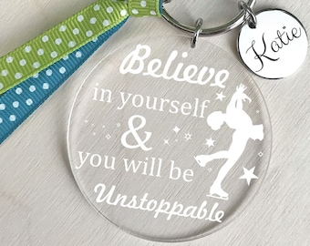 May include: A clear round keychain with the text "Believe in yourself & you will be Unstoppable" and a silhouette of a figure ice skating. The keychain has a green and blue ribbon with white polka dots attached to it. A silver charm with the name "Katie" is attached to the keychain.