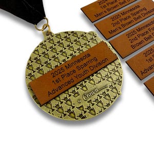 May include: A gold-colored medal with a black ribbon, featuring a brown leather band with text. The medal has a repeating crown pattern. Several brown leather tags with text are also visible. The text includes "2025 Minnesota 1st Place Sparring Advanced Youth Division."