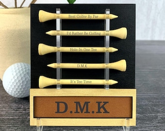 Può includere: Un porta tee da golf in legno con sfondo nero e cinque tee da golf. Il porta tee ha una placca in pelle marrone con il testo "D.M.K" su di essa. I tee da golf hanno il seguente testo inciso su di essi: "Best Golfer By Par", "I'd Rather Be Golfing", "Hole-In-One Tee", "D.M.K", e "It's Tee Time".