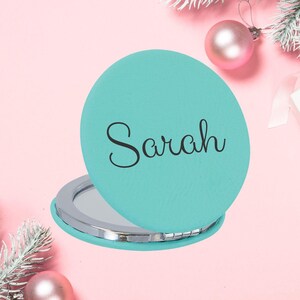 Peut inclure: Un miroir de poche turquoise avec le nom "Sarah" en écriture noire. Le miroir est ouvert, révélant une surface réfléchissante. Le fond présente des ornements roses, une boîte cadeau et des branches de sapin, suggérant un thème de vacances.