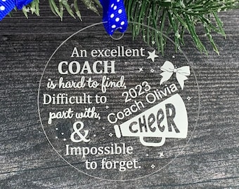 May include: A clear round ornament with a white inscription that reads "An excellent COACH is hard to find, Difficult to part with, & Impossible to forget."  The inscription also includes a megaphone with the word "CHEER" inside and the text "Coach Olivia 2023".