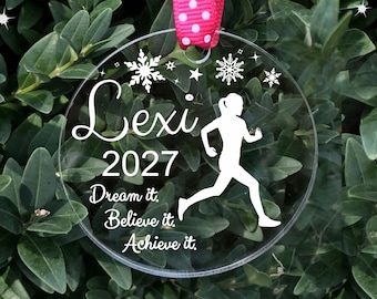 May include: A clear round ornament with a white silhouette of a runner, the text "Lexi 2027 Dream it. Believe it. Achieve it." and snowflakes.