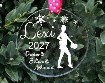 May include: A clear round acrylic ornament with a white silhouette of a cheerleader holding pom-poms. The ornament has the text "Lexi 2027 Dream it. Believe it. Achieve it." printed on it.