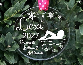 May include: Clear round acrylic ornament with a white silhouette of a swimmer, snowflakes, and stars. The ornament also has the text "Lexi 2027 Dream it. Believe it. Achieve it."