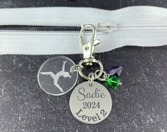 May include: A silver keyring with a clear acrylic charm featuring a gymnast silhouette. The keyring also has a silver charm with the text "Sadie 2024 Level 2" and green and purple beads.
