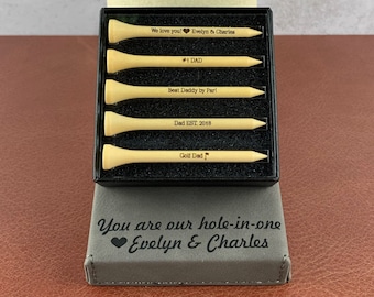 May include: A set of four wooden golf tees in a black box with engraved messages. The messages include "We love you! Evelyn & Charles", "#1 DAD", "Best Daddy by Par!", "Dad EST. 2018", and "Golf Dad". The box is sitting on a brown surface. The box also has a gray lid with the text "You are our hole-in-one Evelyn & Charles" engraved on it.