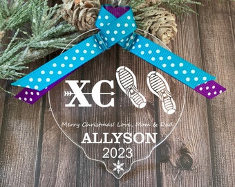 Puede incluir: Un adorno navideño de acrílico transparente con un diseño de copo de nieve blanco. El adorno presenta un diseño de carrera de campo a través blanco con el texto "XC" y dos zapatillas para correr. El adorno también incluye el texto "¡Feliz Navidad! Con amor, Mamá y Papá" y "ALLYSON 2023". El adorno está decorado con una cinta de lunares azul y morado.