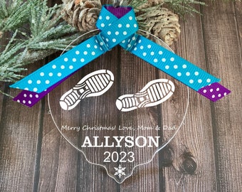 Könnte beinhalten: Ein transparenter Weihnachtsbaumschmuck aus Acryl mit einem Schneeflockenmuster. Er zeigt zwei weiße Fußabdrücke und den Text "Merry Christmas! Love, Mom & Dad" und "ALLYSON 2023". Der Schmuck ist mit einem blau-violetten gepunkteten Band verziert.