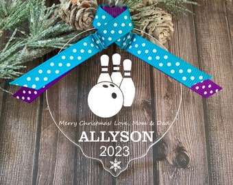 May include: A clear acrylic Christmas ornament with a white bowling ball and pins design. The ornament is decorated with a blue and purple polka dot ribbon and has the text "Merry Christmas! Love, Mom & Dad" and "ALLYSON 2023" printed on it.