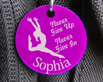 May include: A circular purple tag featuring a gymnast silhouette and the words "Never Give Up, Never Give In" and the name "Sophia" in white. It is attached to a silver clip.