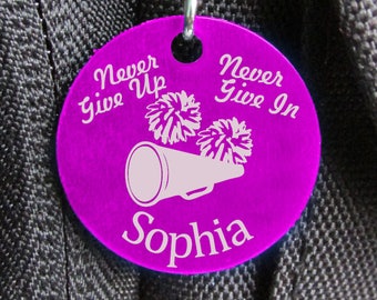Peut inclure: Porte-clés rond violet avec le texte "Never Give Up" et "Never Give In" avec un mégaphone blanc et des pompons de majorette. Le nom "Sophia" est écrit sous le mégaphone.