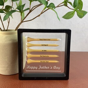 May include: A black framed picture with a clear background. Inside the frame are five golf tees with engraved messages. The messages read "Happy Father's Day!", "Golf Papa", "#1 PAPA", "Best Papa by Par", and "Nice shot, Papa!" The bottom of the frame reads "Happy Father's Day".