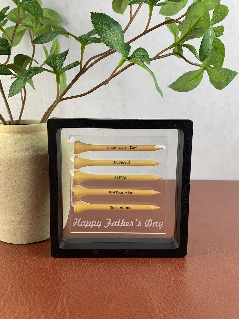 May include: A black framed picture with a clear background. Inside the frame are five golf tees with engraved messages. The messages read "Happy Father's Day!", "Golf Papa", "#1 PAPA", "Best Papa by Par", and "Nice shot, Papa!" The bottom of the frame reads "Happy Father's Day".
