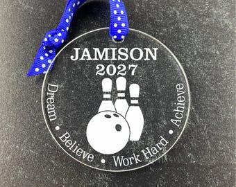 Puede incluir: Un adorno redondo transparente con un diseño de bola de bolos y bolos blancos. El texto "JAMISON 2027" está en la parte superior del adorno. El texto "Dream Believe Work Hard Achieve" está alrededor del borde del adorno.