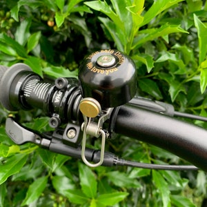 May include: A black bicycle bell with a gold-colored inscription that reads "Ride safe Dear, We love you". The bell is mounted on a black bicycle handlebar.