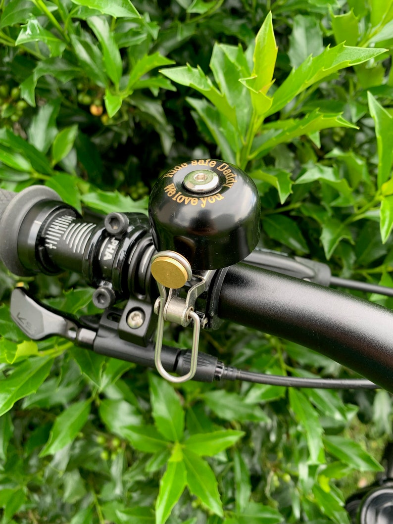 May include: A black bicycle bell with a gold-colored inscription that reads "Ride safe Dear, We love you". The bell is mounted on a black bicycle handlebar.