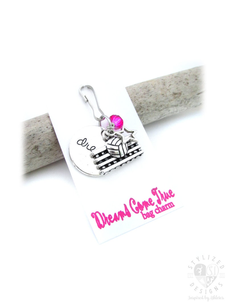 May include: A silver charm with a volleyball, a star, and a heart with the word "dream" on it. The charm is attached to a silver clip. The charm is on a white card with the words "Dreams Come True bag charm" in pink.