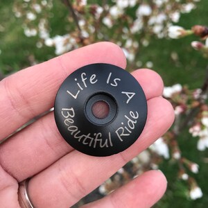 May include: Black round bike stem cap with the words "Life is a Beautiful Ride" engraved in silver lettering.