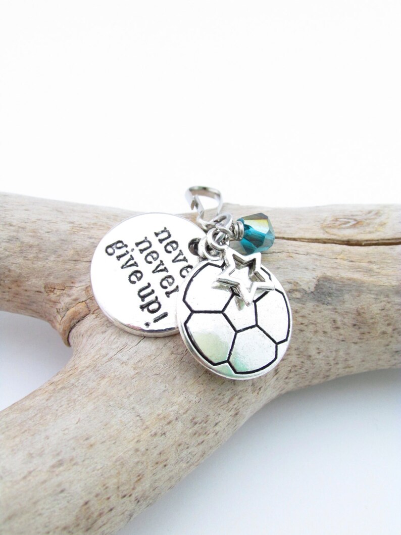 May include: Silver charm with a soccer ball, a star, and a blue crystal. The charm has the words "never give up!" engraved on it.