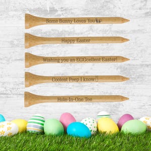 May include: Five wooden golf tees with Easter-themed messages engraved on them. The messages are "Some Bunny Loves You", "Happy Easter", "Wishing you an EGGcellent Easter!", "Coolest Peep I know", and "Hole-In-One Tee". The tees are arranged vertically on a white wooden background. There are colorful Easter eggs in the grass at the bottom of the image.