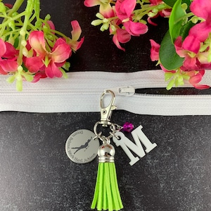 May include: A silver keychain with a green tassel, a silver charm with the text "Dream. Believe. Work Hard. Achieve." and a silhouette of a gymnast, and a white letter "M".