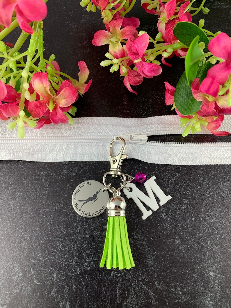 May include: A silver keychain with a green tassel, a silver charm with the text "Dream. Believe. Work Hard. Achieve." and a silhouette of a gymnast, and a white letter "M".