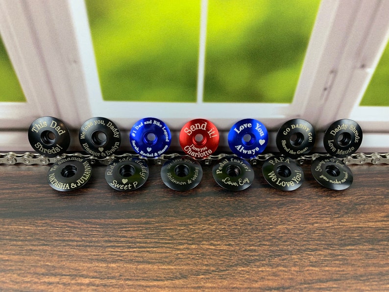 May include: Ten personalized bike stem caps with various colors and engraved messages. The caps are black, blue, red, and silver. The messages include "This Dad Shreds", "We love you, Daddy", "#1 Dad and Bike Legend", "Send it! I love you Charlotte", "Love You Always", "Go Daddy Go! Shred the Gear", "Daddy Shreds Love Maude", "We love you Chiannon & Wesley", "Pedal faster Love Evy", "Papa We Love You!"