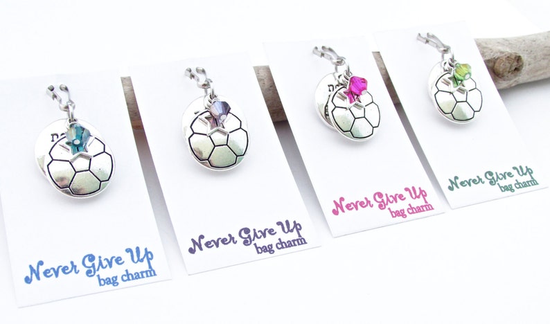 May include: Four silver soccer ball bag charms with different colored crystals. Each charm has the text "Never Give Up" and "bag charm" printed on a white card.